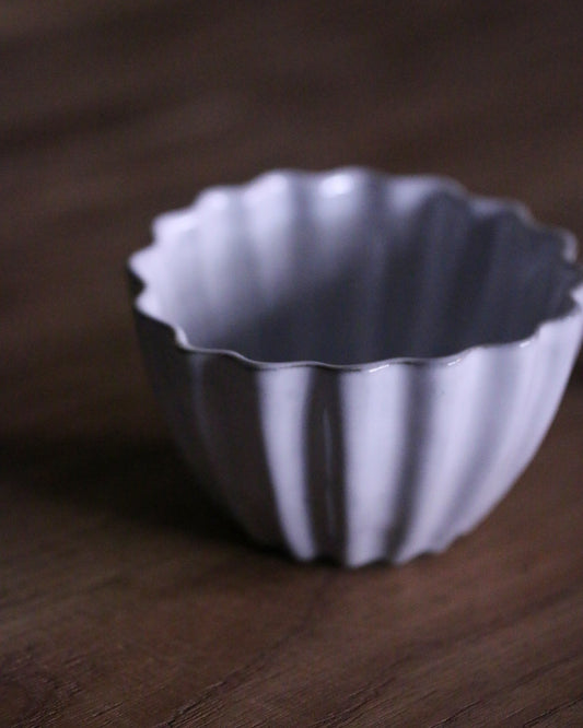 Pale lilac cup with deep fluted ribs and a softly scalloped rim, on a dark wooden surface.
