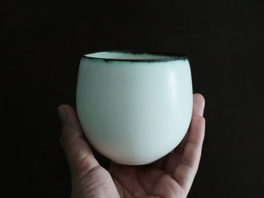 Rounded celadon cup in soft blue-green with a dark rim, held in a hand against black.
