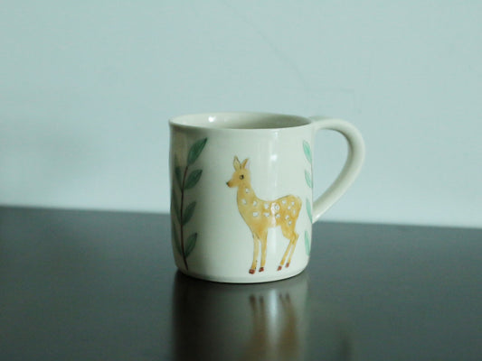 White mug painted with a spotted fawn among green leaves, with a slim loop handle.