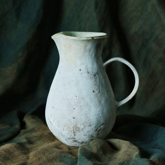 Tall white speckled jug with a narrow neck and a slim loop handle, against green cloth.