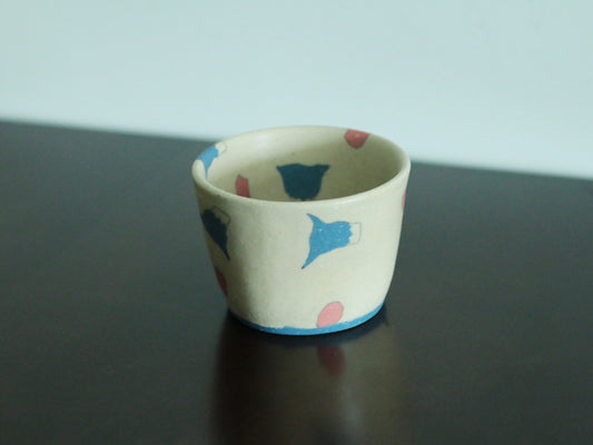 Cream cup painted with small blue and red shapes, standing on a dark reflective surface.