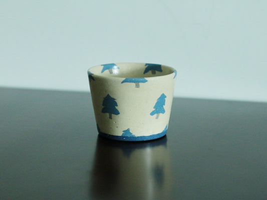 Cream cup painted with a row of small blue trees, on a dark reflective surface.