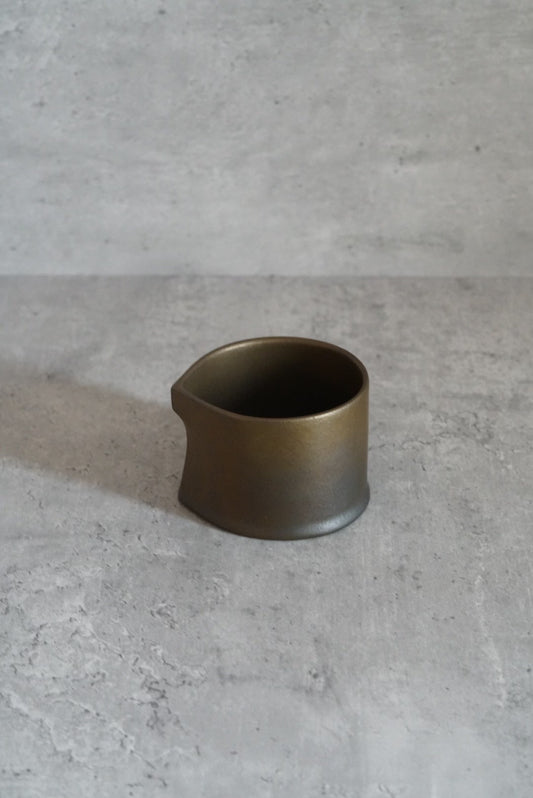 Small cup in dark bronze metallic glaze with a stepped base, on grey concrete.