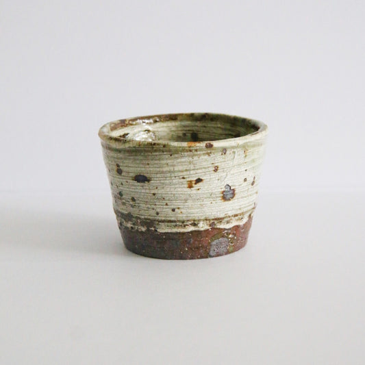 Straight-sided cup in speckled cream slip over a band of bare rust-brown clay at the foot.