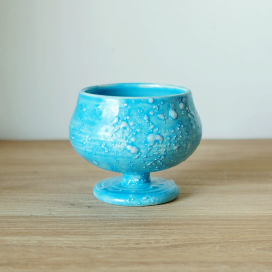 Stemmed cup in vivid turquoise, its glaze bubbled and crawled into pale spots, on a wooden table.