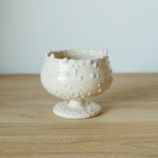 Cream stemmed cup with a crusty beaded surface and a softly uneven rim, on a wooden table.