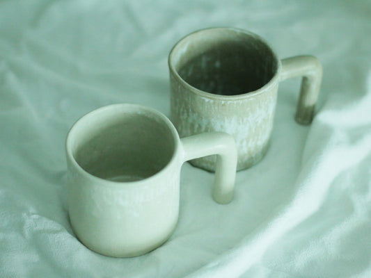 Two pale green mugs with squared handles and softly pitted surfaces, on rumpled white cloth.