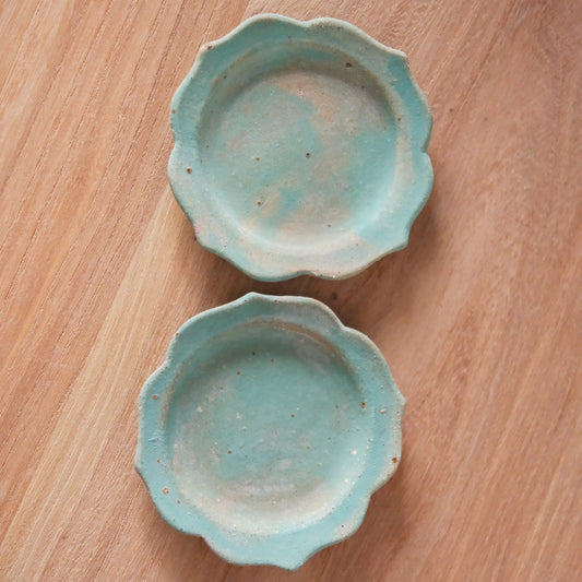 Two small pale green dishes with softly scalloped flower rims, on a light wooden table.