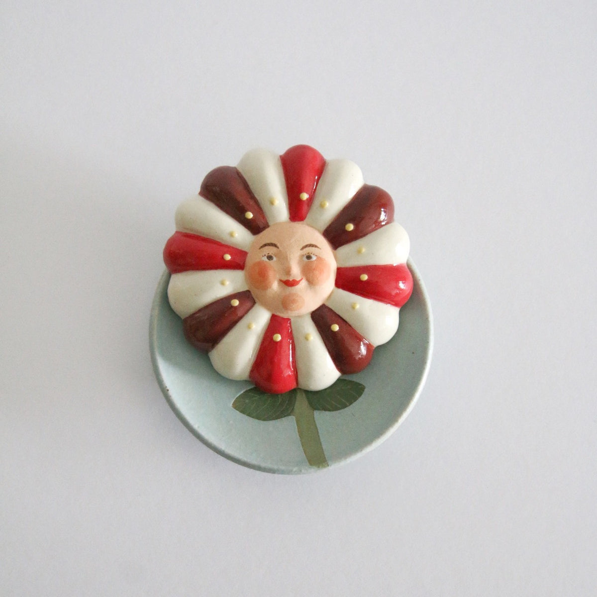 Ceramic flower with red and white petals and a painted face at its centre, on a pale green dish.