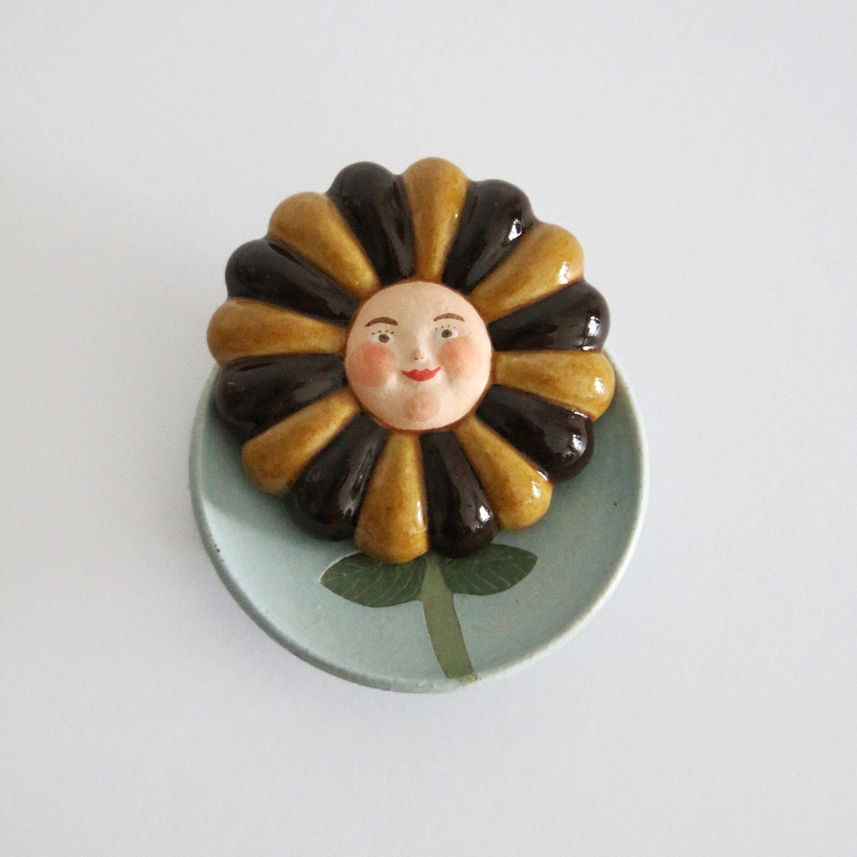 Ceramic flower with alternating mustard and black petals and a painted face, on a pale green dish.