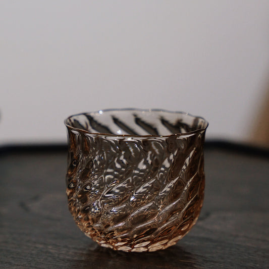 Warm brown glass tea cup with a twisted, scale-like ribbed surface, on dark wood.