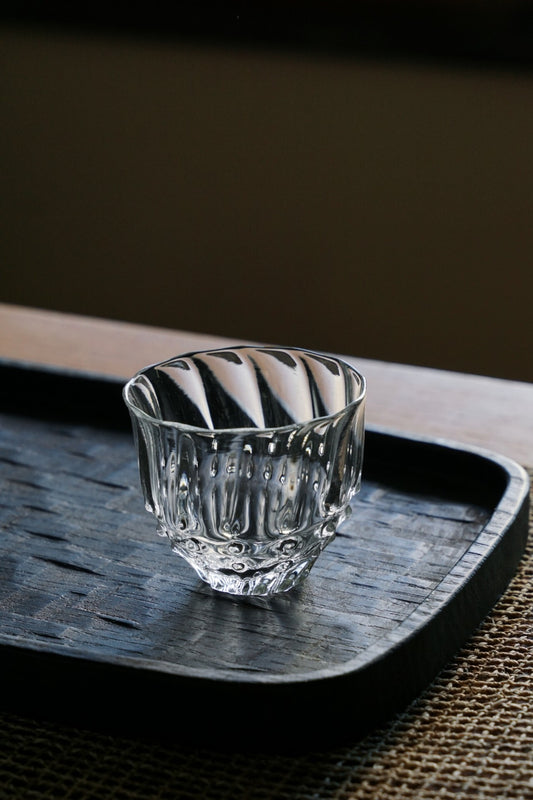 Small clear glass tea cup with softly rippled sides, sitting on a dark lacquered tray.