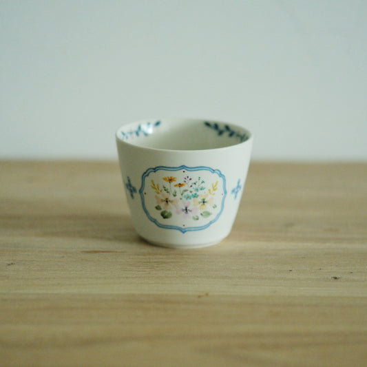 White cup painted with a small framed posy of blue and yellow flowers, on a wooden table.
