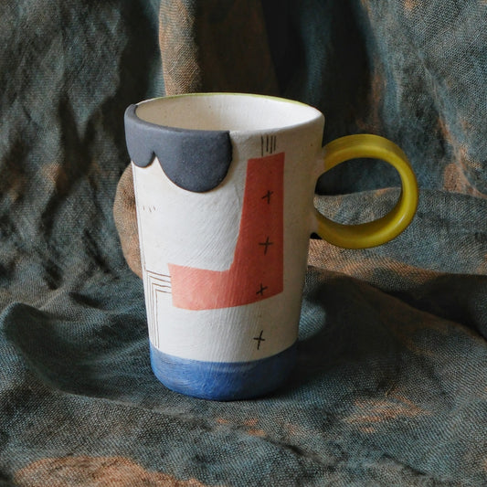 Tall cream mug painted in bold red, navy and grey blocks, with a bright yellow ring handle.