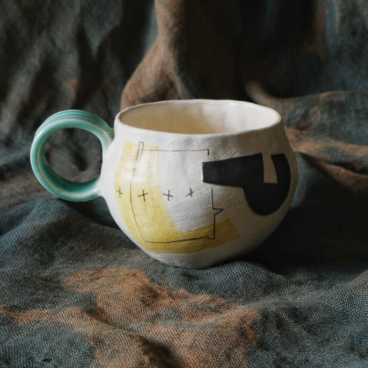 Wide cream mug painted with black and yellow shapes, with a pale green ring handle.