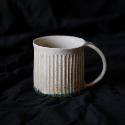 Cream mug carved with close vertical lines, a green glaze pooling at its base, on black cloth.