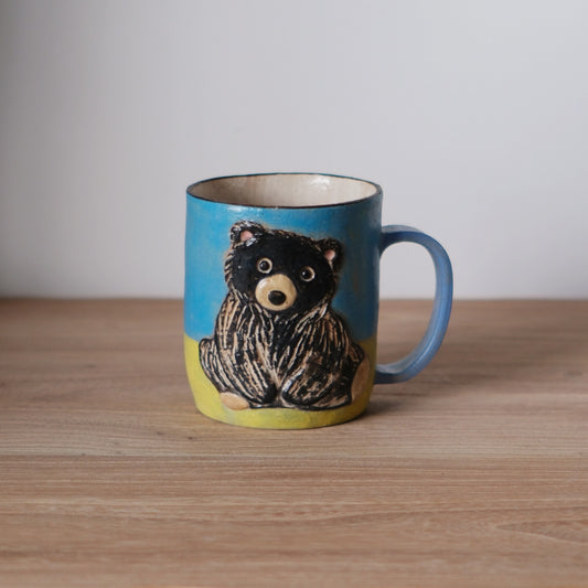 Blue mug painted with a black bear standing in yellow grass, with a matching blue handle.