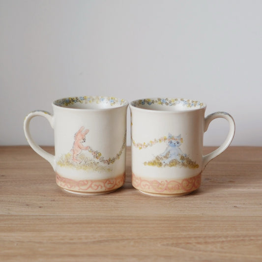 Two white mugs painted with small animals in a flowering meadow above a soft pink band.