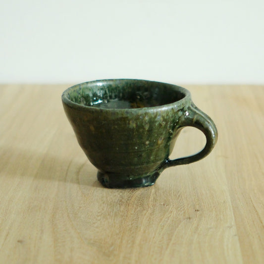 Deep green glossy mug on a small foot with a chunky loop handle, on a light wooden table.