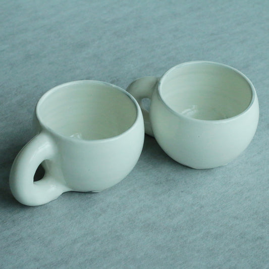 Two pale mint green mugs with rounded bodies and small ring handles, on soft blue cloth.