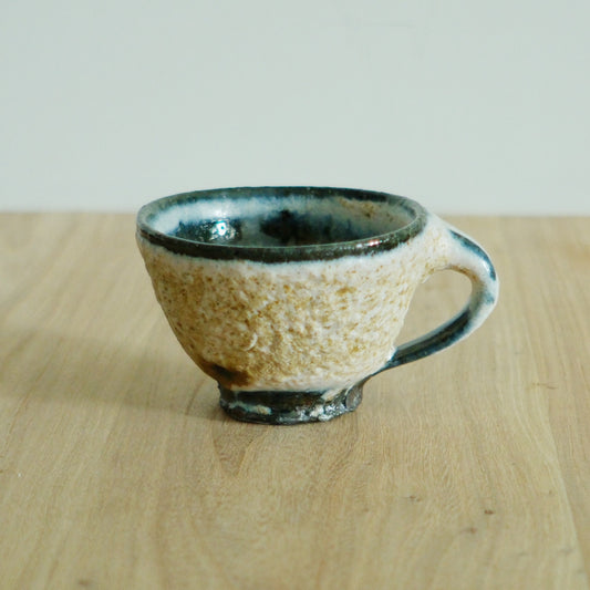 Wide cream mug speckled with iron, its rim and handle washed with deep blue-green glaze.
