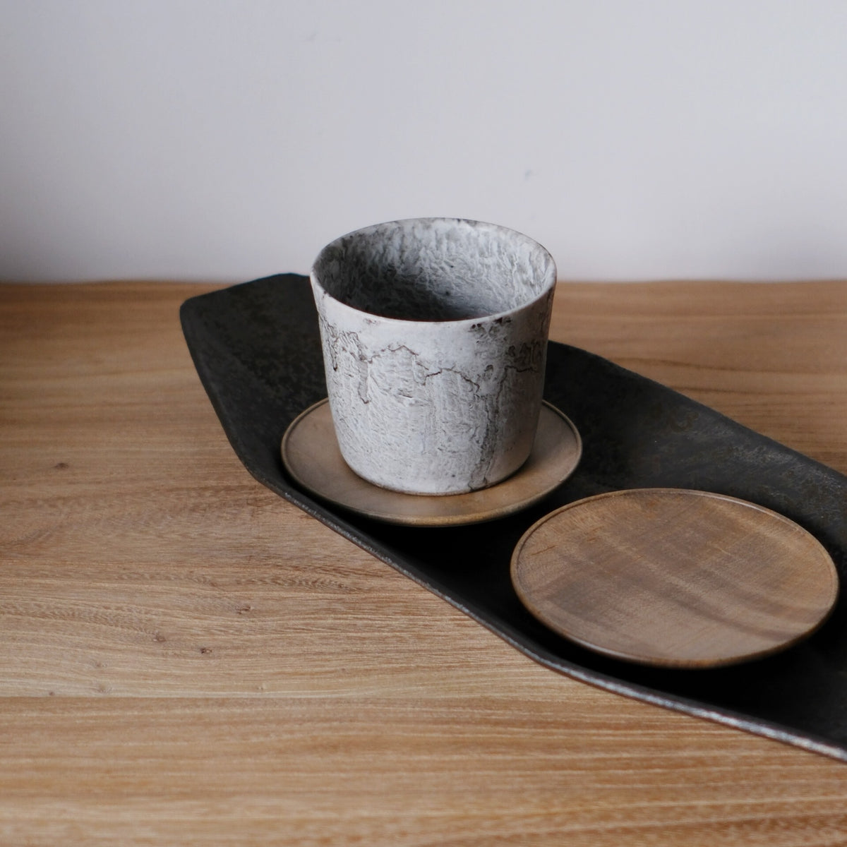 Round wooden saucer beside a speckled grey cup on a dark board, on a light wooden table.