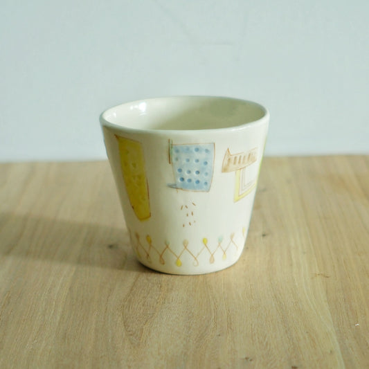 Cream cup painted with loose yellow blocks and small blue stitch marks, on a wooden table.