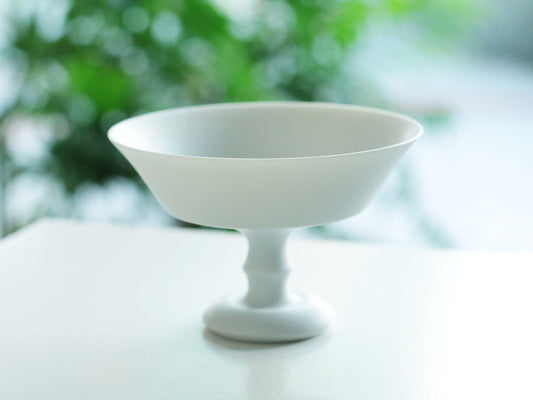 White porcelain dessert cup with a wide shallow bowl on a slender turned stem, against a bright window.