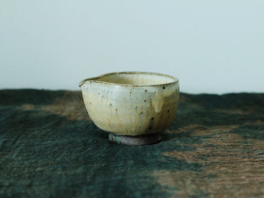 Small footed pouring bowl in speckled cream slip with a drawn spout, on a dark weathered board.
