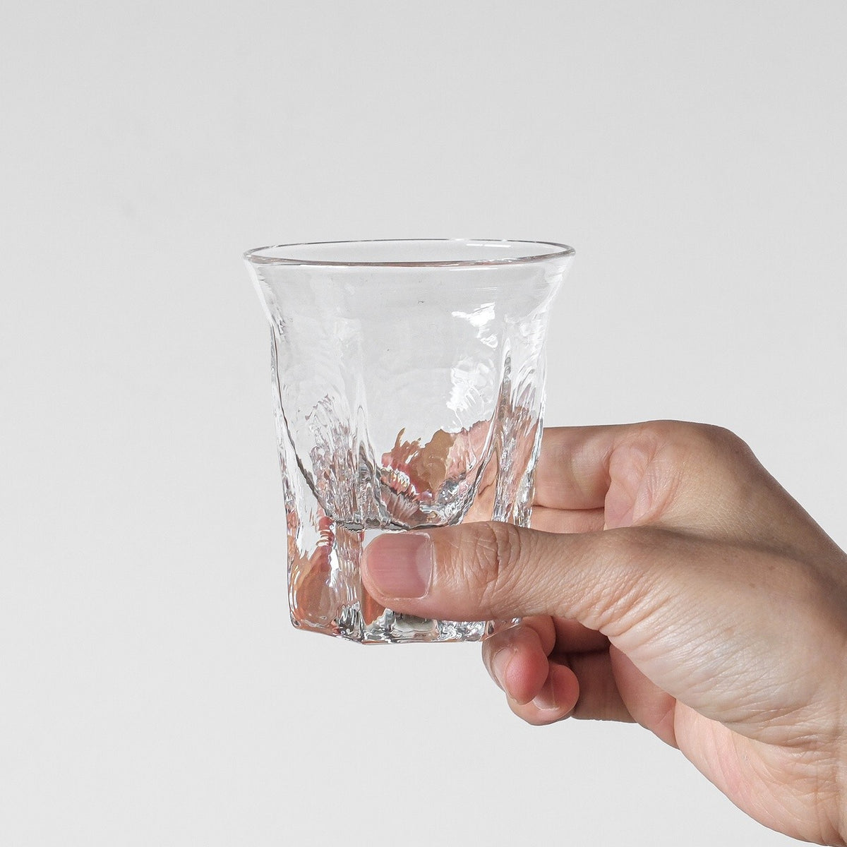 Clear glass cup with an irregular hammered surface, held in a hand against a pale ground.