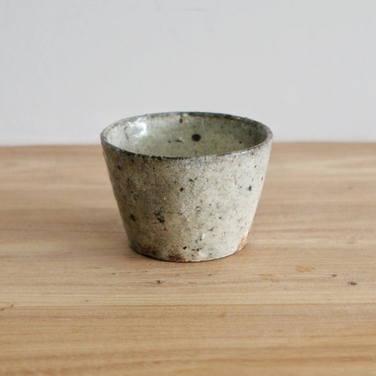 Tapered wood-fired cup in pale grey with a coarse sandy texture, on a light wooden table.