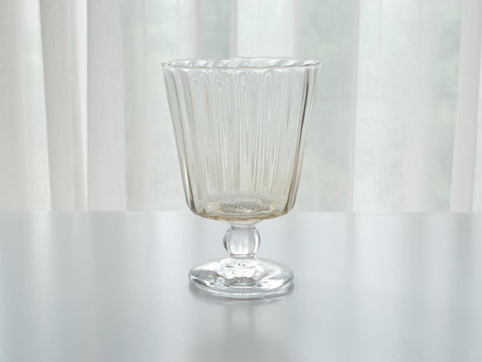 Stemmed glass with fine vertical ribbing on a short round foot, against a soft white curtain.