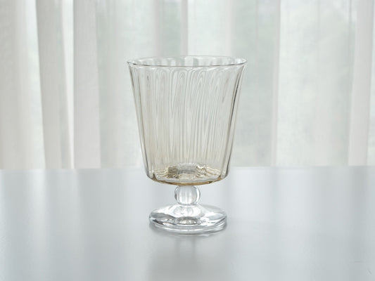 Ribbed stemmed glass on a short foot, slightly shorter in the bowl, against a soft white curtain.