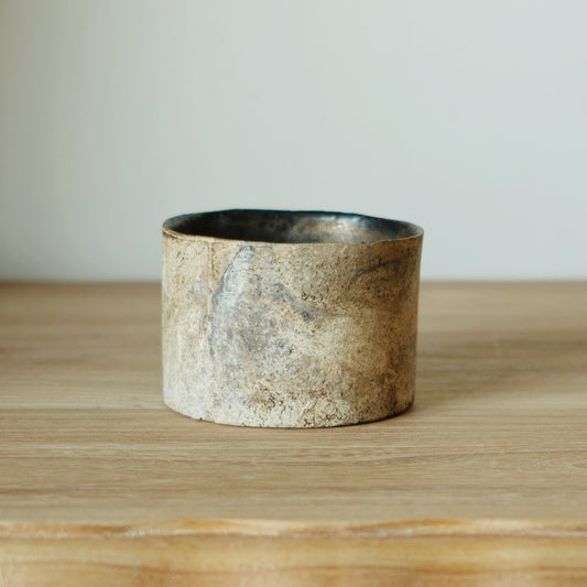 Straight-sided cup in rough mottled cream and grey with a dark glazed interior, on a wooden shelf.