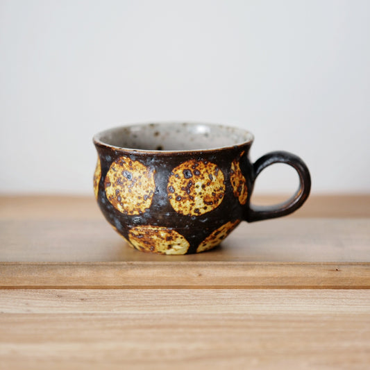 Dark mug ringed with large golden-yellow spots, with a chunky loop handle, on a wooden shelf.