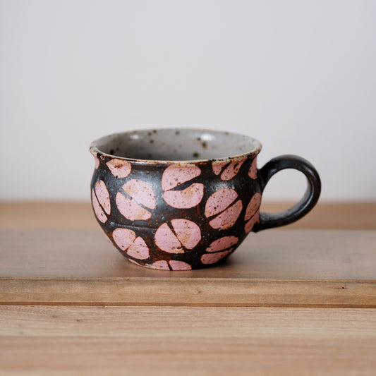 Dark mug ringed with soft pink spots and a loop handle, on a light wooden shelf.