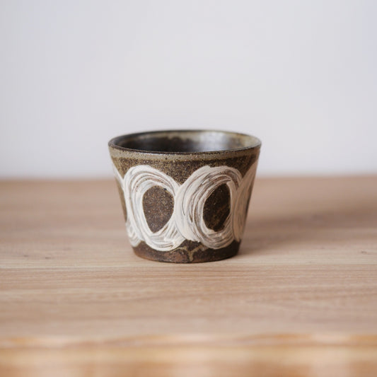 Tapered brown cup painted with linked white rings, on a light wooden shelf.