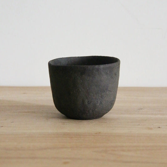 Small deep black cup with a rough matte surface and a slightly flared rim, on wood.
