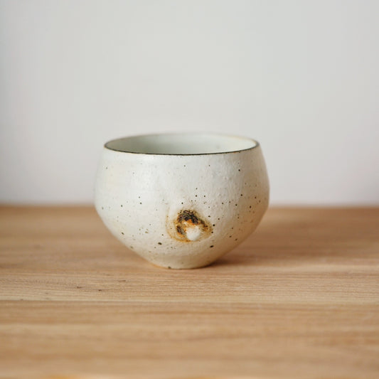 Rounded cream cup speckled with dark grit, a small amber glaze pool low on one side.