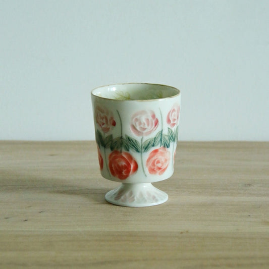 White stemmed cup painted with full pink roses on green stems, on a light wooden table.