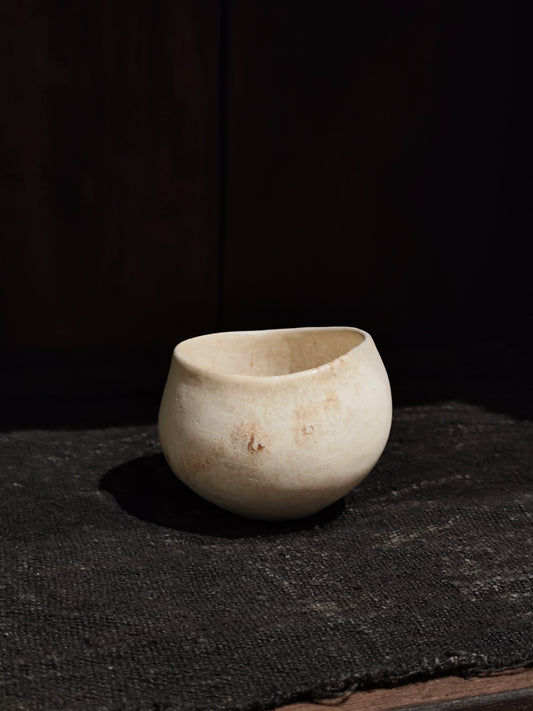 Small pale cream cup marked with soft rust blushes, resting on dark textured cloth.