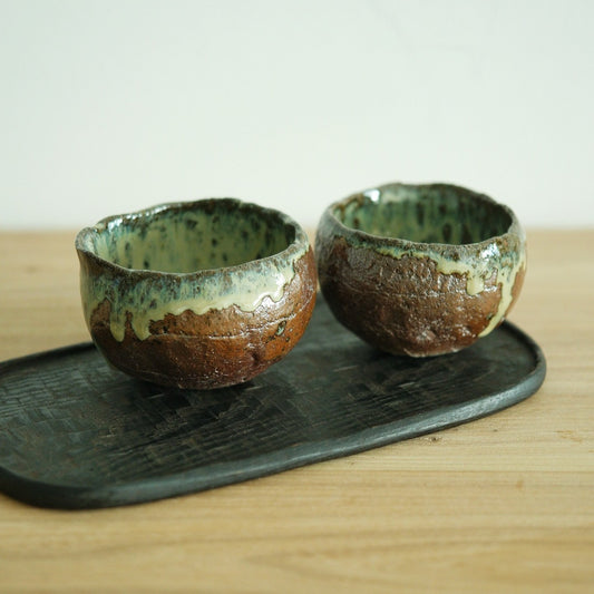 Two cups with green ash glaze running over bare rust-brown clay, on a dark board.