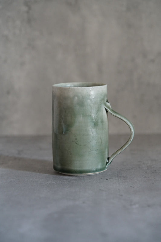 Tall celadon mug in soft blue-green with a slim wire-thin handle, on grey concrete.