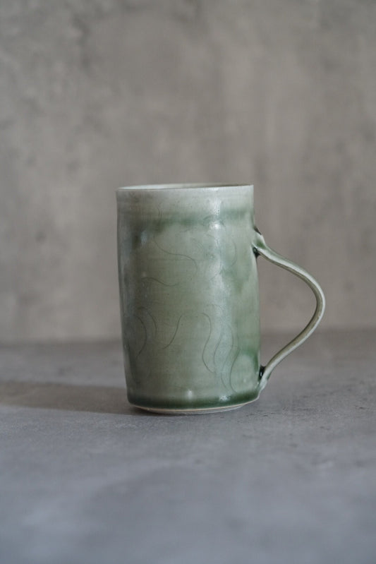 Tall celadon mug in pale green with a very fine looping handle, on grey concrete.