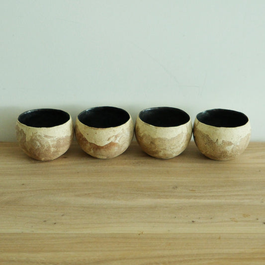 Four cream cups with rough torn surfaces and black glazed interiors, on a pale wooden shelf.