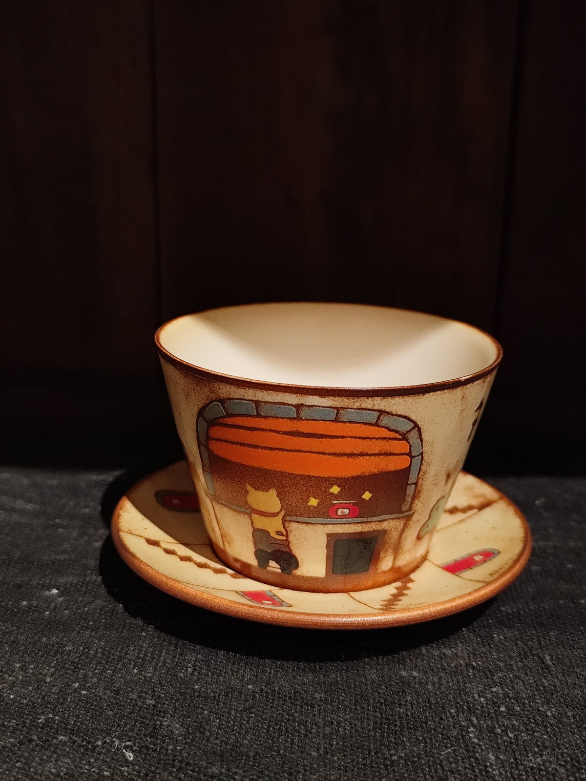 Cream cup and saucer with brown rims, painted with a glowing orange arched oven set into a wall.