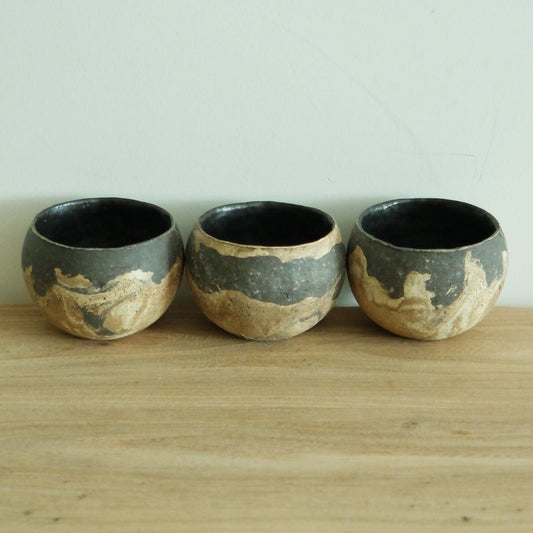 Three round cups banded in cream and slate grey with black interiors, on a wooden shelf.