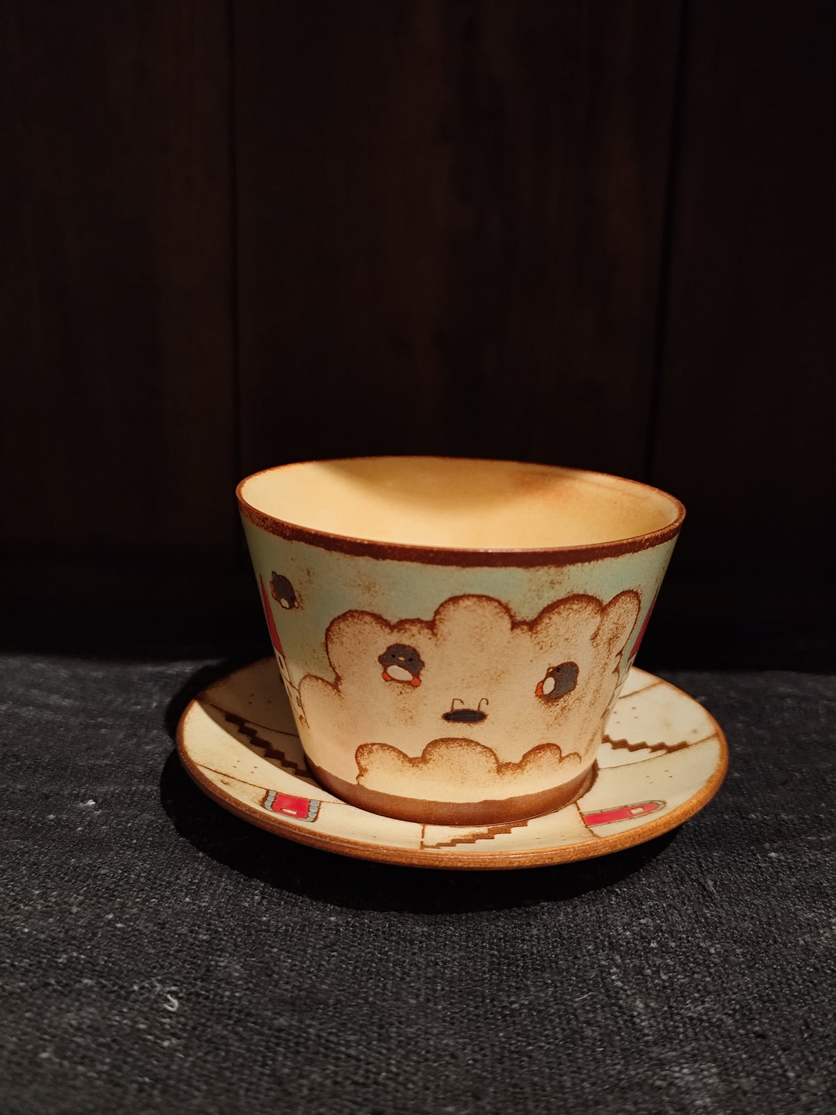 Cream cup and saucer with brown rims, painted with a soft cloud shape wearing a small face.