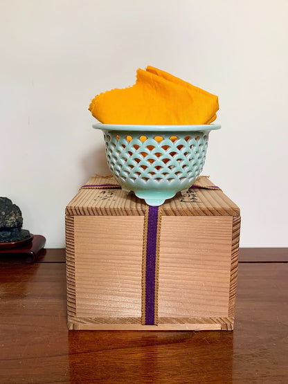 A light blue glazed ceramic strainer with a diamond lattice sits atop a wooden box tied with purple ribbon.