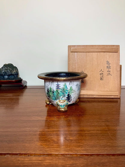 A glazed ceramic pot with a forest scene, three feet, and a dark rim.